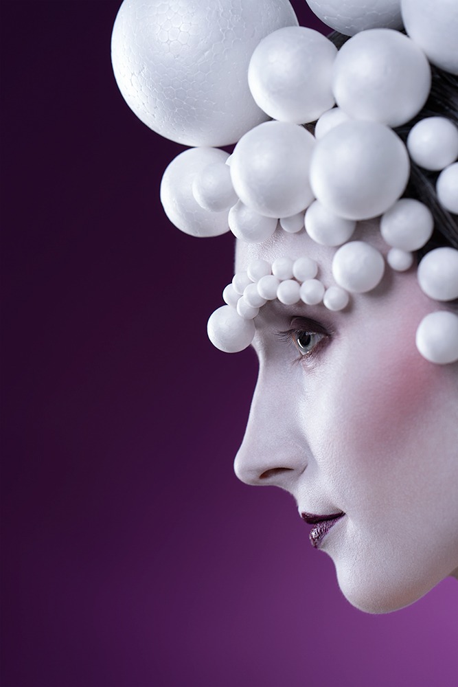 Profil kobiety z białym makijażem i nakryciem głowy z białych kul na fioletowym tle.