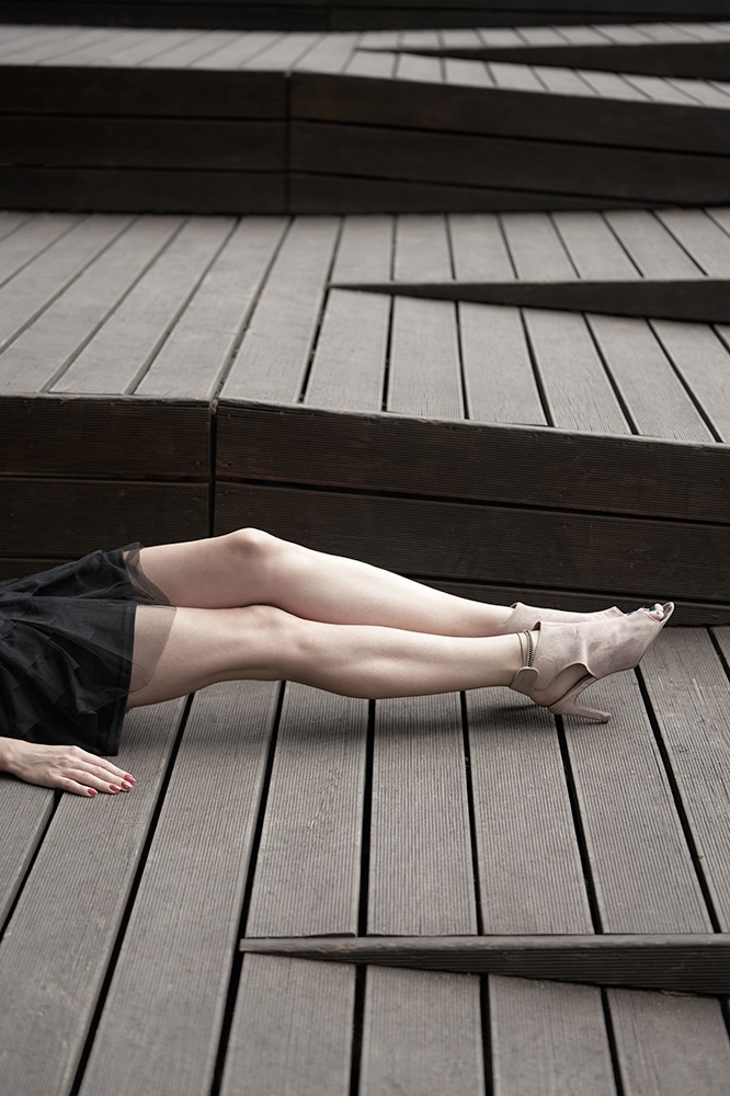 Kobieta w czarnej tiulowej spódnicy i beżowych szpilkach leży na drewnianych schodach, paznokcie pomalowane na czerwono.