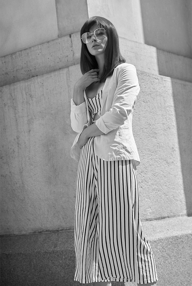 Czarno-białe zdjęcie kobiety w okularach, w paski kombinezonie i białej marynarce, opierającej się o betonową ścianę w słoneczny dzień.