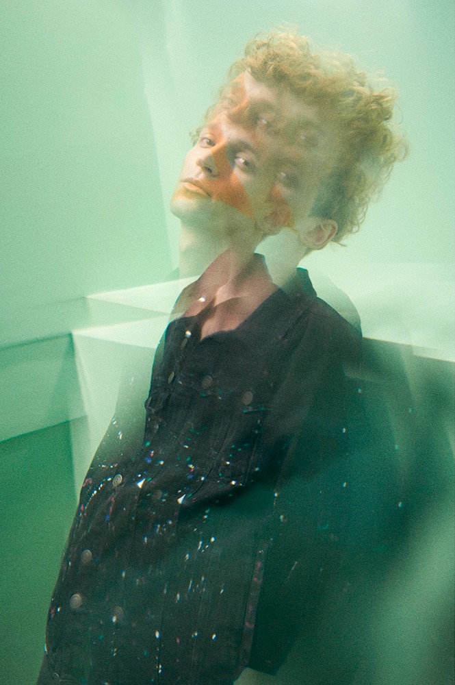 Podwójna ekspozycja portretu młodego mężczyzny w studio w Warszawie, z pomarańczowym wzorem na twarzy i kurtką pokrytą kolorowymi plamami farby, na tle zielonych ścian.