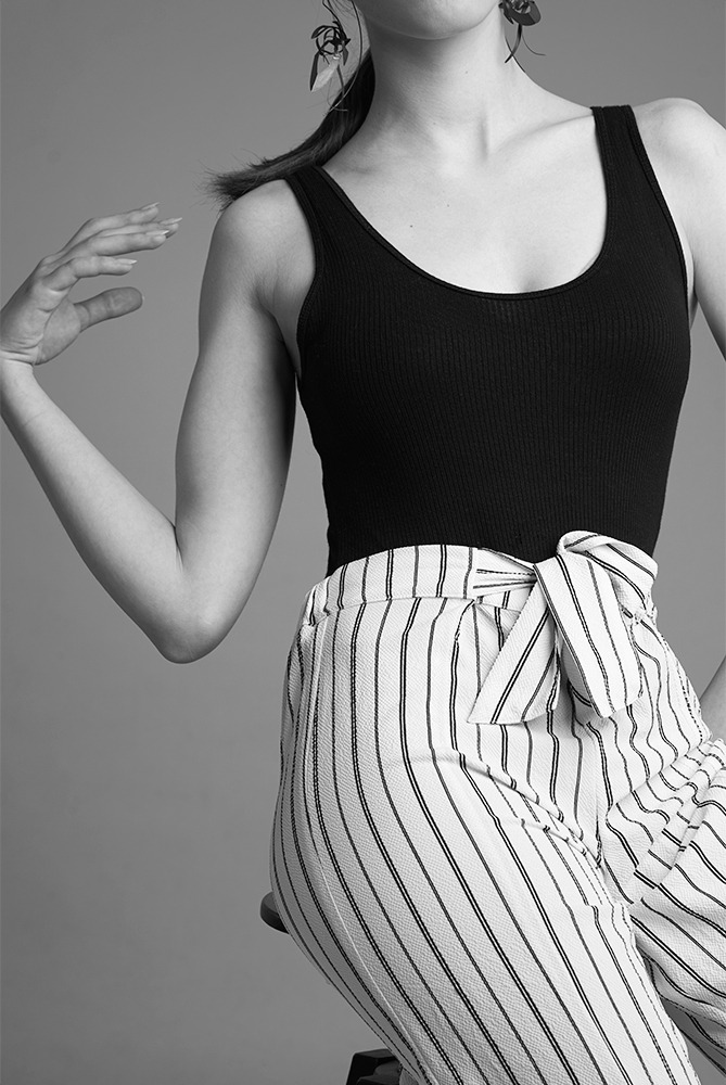 Czarno-białe zdjęcie modelki w czarnym topie i spodniach w paski z kokardą, pozującej na szarym tle z wyrazistymi kolczykami.