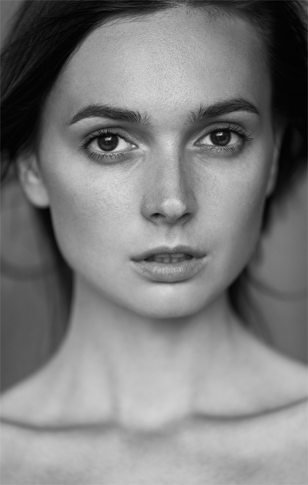 Portret młodej kobiety w czerni i bieli, z delikatnym makijażem i naturalnym wyrazem twarzy, włosy lekko rozwiane, oświetlenie podkreśla rysy twarzy.