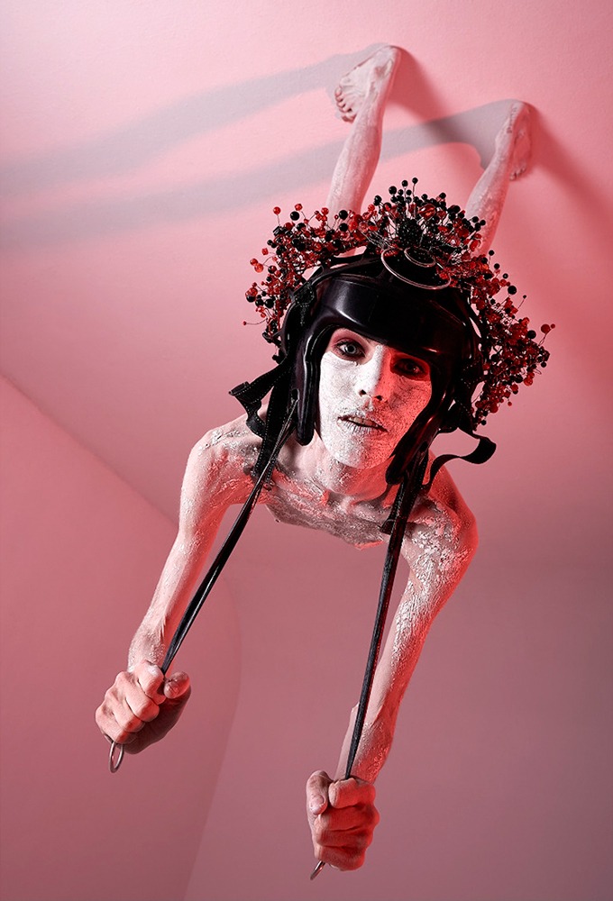 Osoba z pomalowaną na biało skórą, w czarnym hełmie i ozdobnym wieńcu, trzymająca w dłoniach metalowe pręty, z nogami wystającymi z sufitu w czerwonym oświetleniu.