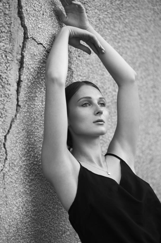 Czarno-białe zdjęcie kobiety w czarnym topie, opierającej się o szorstką ścianę z uniesionymi rękami, delikatny naszyjnik z wisiorkiem w kształcie serca.