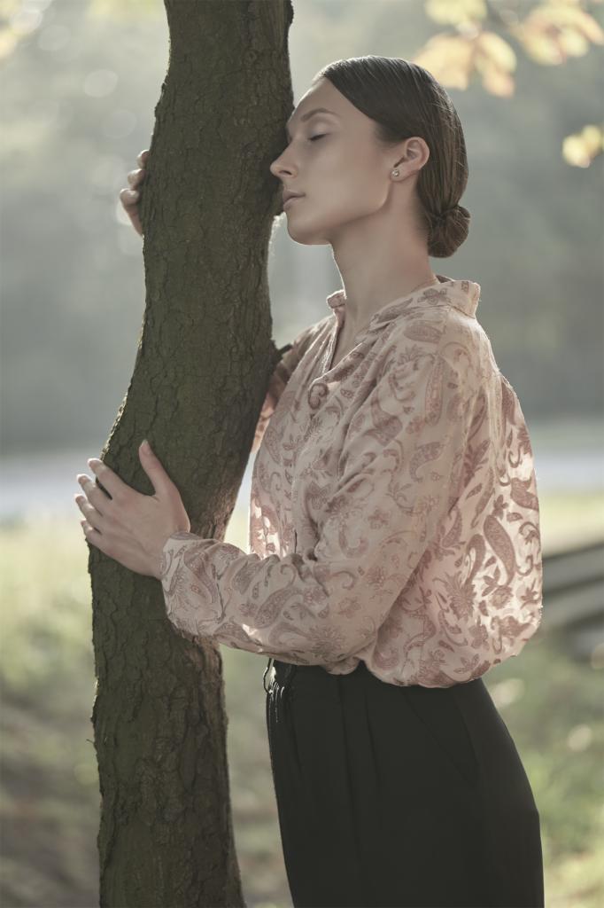 Portret kobiety w koszuli w roślinny wzór, opierającej się o drzewo w parku, z zamkniętymi oczami i subtelnym uśmiechem.