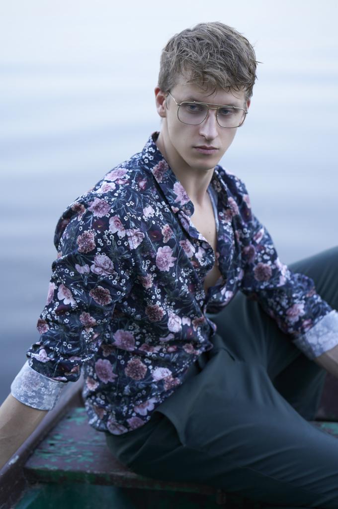 Młody mężczyzna w okularach i kwiecistej koszuli siedzi w łodzi, spoglądając w dół.
