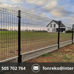 FONREKO - Czarne ogrodzenie panelowe z podmurówką o fakturze kamienia na tle zielonego pola i domu. Słupki ogrodzeniowe z kwadratowymi zakończeniami.