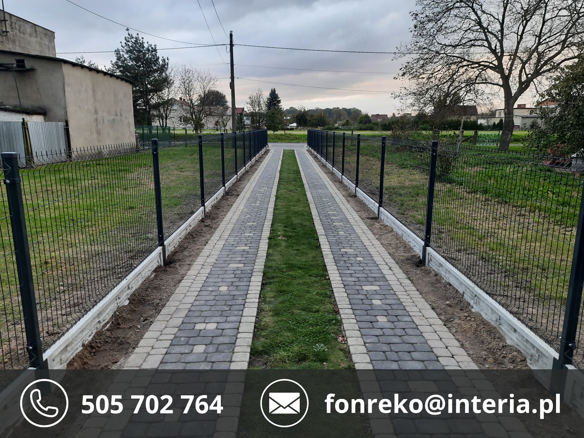 Czarny panelowy płot z podmurówką otacza ścieżkę z kostki. Centralnie ujęta perspektywa, widok na posesję z zielenią i budynkami w tle. Pochmurne niebo.