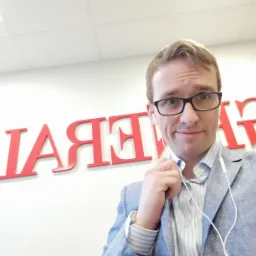 Mężczyzna w okularach i marynarce na tle czerwonego logo firmy ubezpieczeniowej.