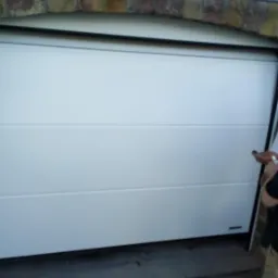 Biała, segmentowa brama garażowa z poziomymi przetłoczeniami, zamontowana w murowanym otworze w Jadownikach. Widoczny fragment osoby obsługującej bramę.