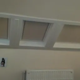 Trzy okna dachowe z roletami w jasnych ramach, zamontowane w poddaszu, widoczne z dołu, z grzejnikiem i obrazkiem na ścianie poniżej.