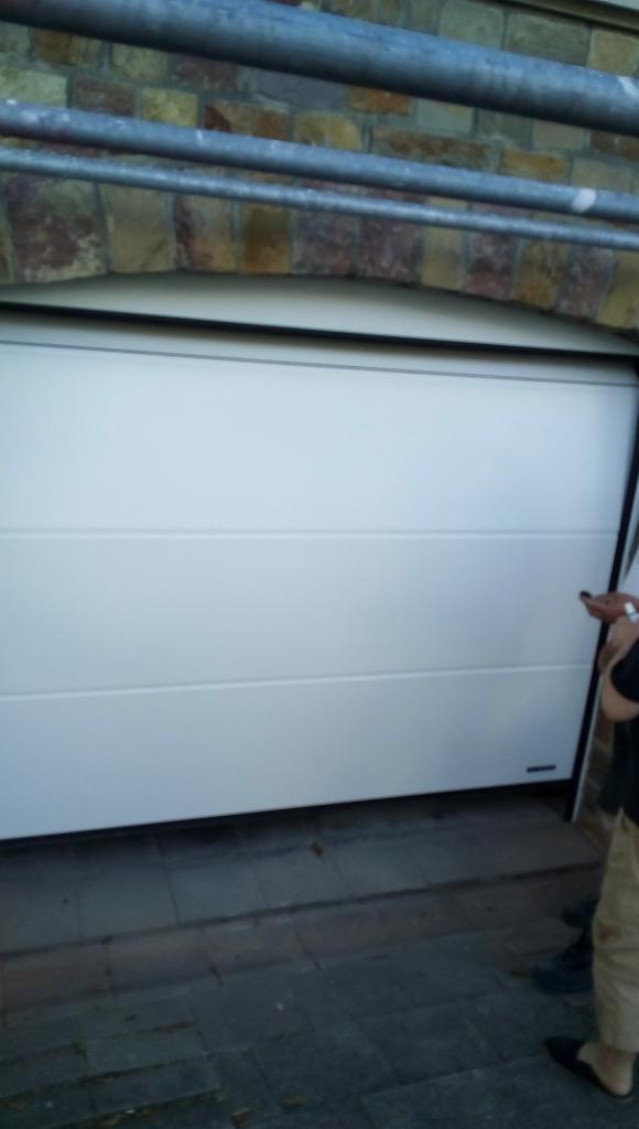 Biała, segmentowa brama garażowa z poziomymi przetłoczeniami, zamontowana w murowanym otworze w Jadownikach. Widoczny fragment osoby obsługującej bramę.