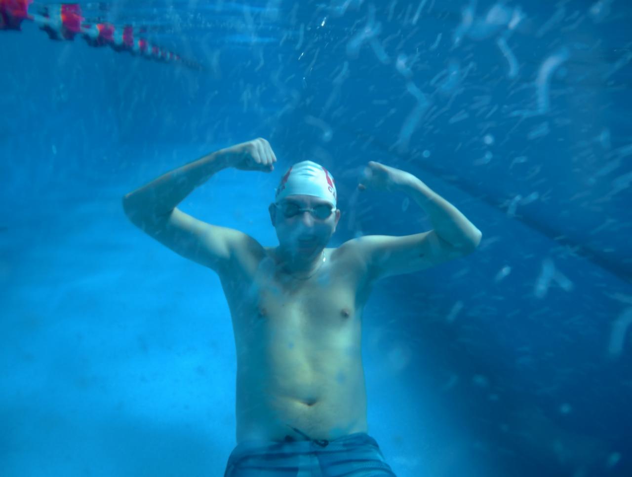 Mężczyzna w czepku i okularach pływackich, widziany pod wodą w basenie, unosi ręce w geście triumfu, widoczne bąbelki powietrza.