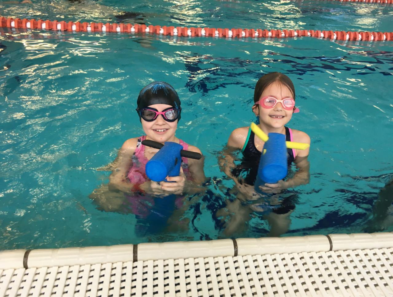 Dwie uśmiechnięte dziewczynki w okularach pływackich trzymają w dłoniach niebieskie makarony do pływania na tle basenu z kafelkami i linami oddzielającymi tory.
