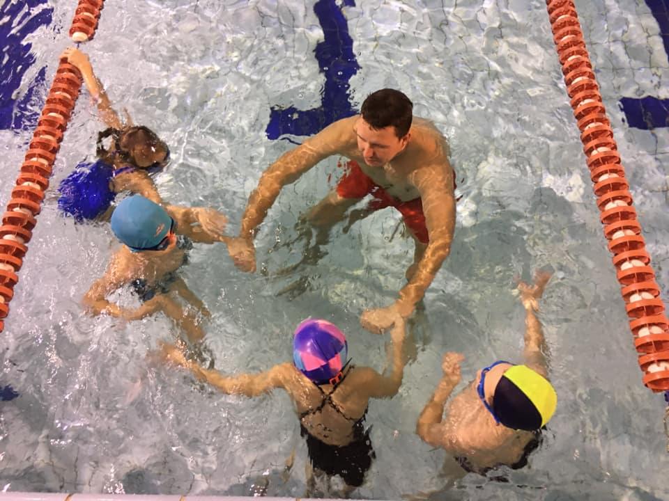 Instruktor w czerwonych spodenkach otoczony czwórką dzieci w czepkach w basenie, trzymających się za ręce, widok z góry.