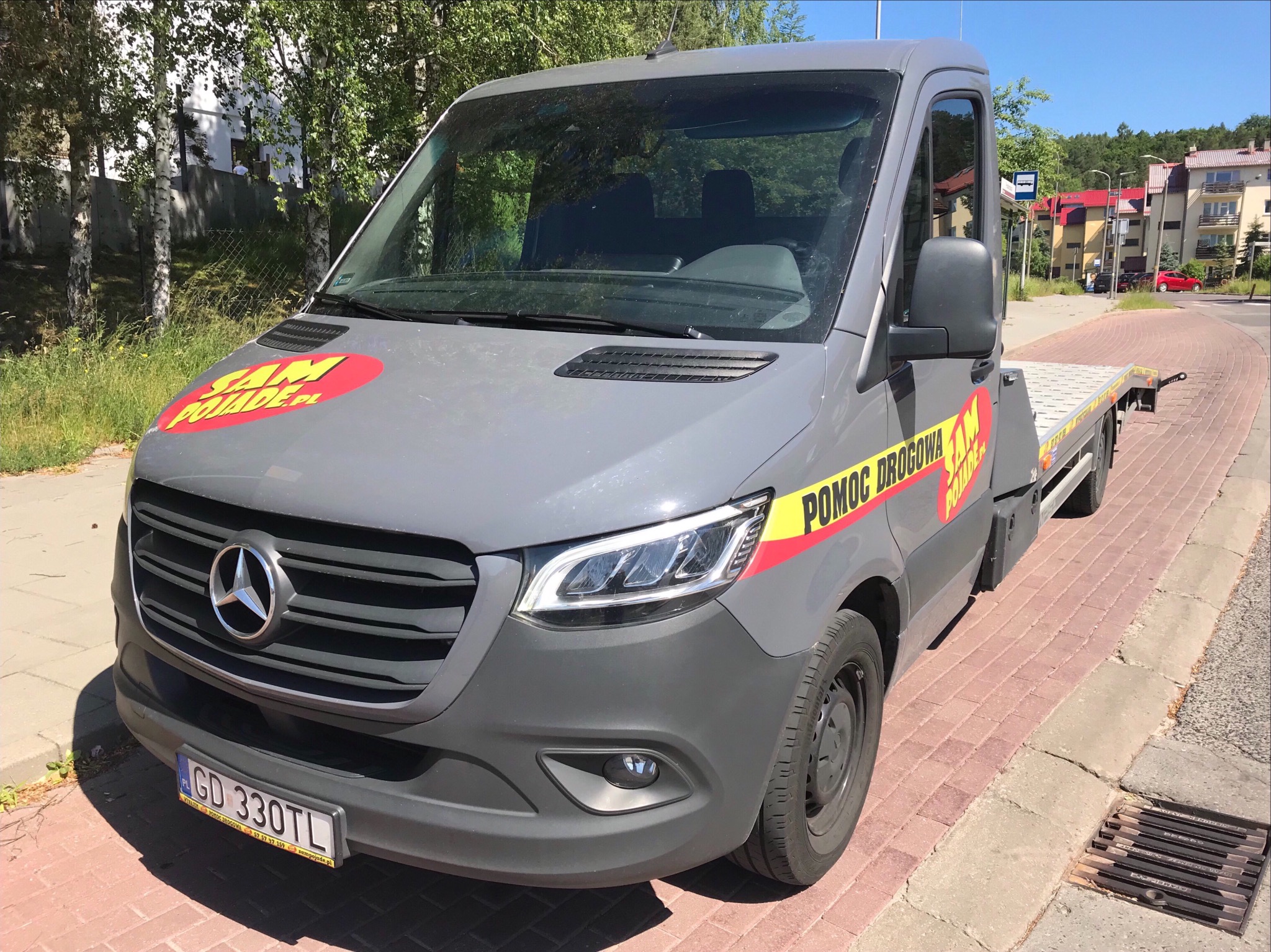 Szary samochód pomocy drogowej marki Mercedes z platformą załadunkową, zaparkowany na chodniku z kostki brukowej, z widocznym logo firmy 'SAM POJADE.PL' i napisem 'POMOC DROGOWA'.