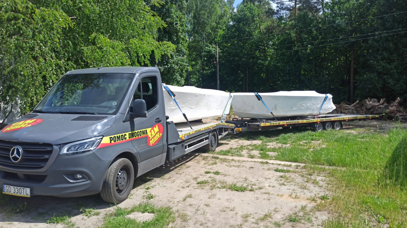 Szara laweta z napisem 'Pomoc Drogowa' przewozi dwie owinięte folią łodzie na przyczepie w słoneczny dzień.