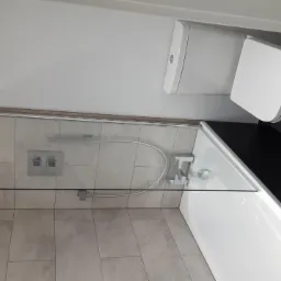 Nowoczesna łazienka po remoncie: beżowe płytki na ścianach, szklany parawan nawannowy z chromowanymi elementami, biała wanna i toaleta, ciemnoszara podłoga z płytek.