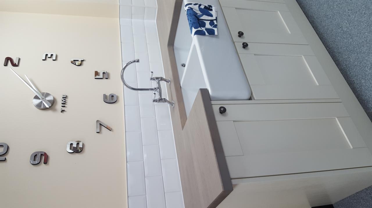 Biała umywalka wbudowana w szafkę z blatem w odcieniach szarości, bateria z kurkami, białe kafelki na ścianie i zegar z naklejanymi cyframi.