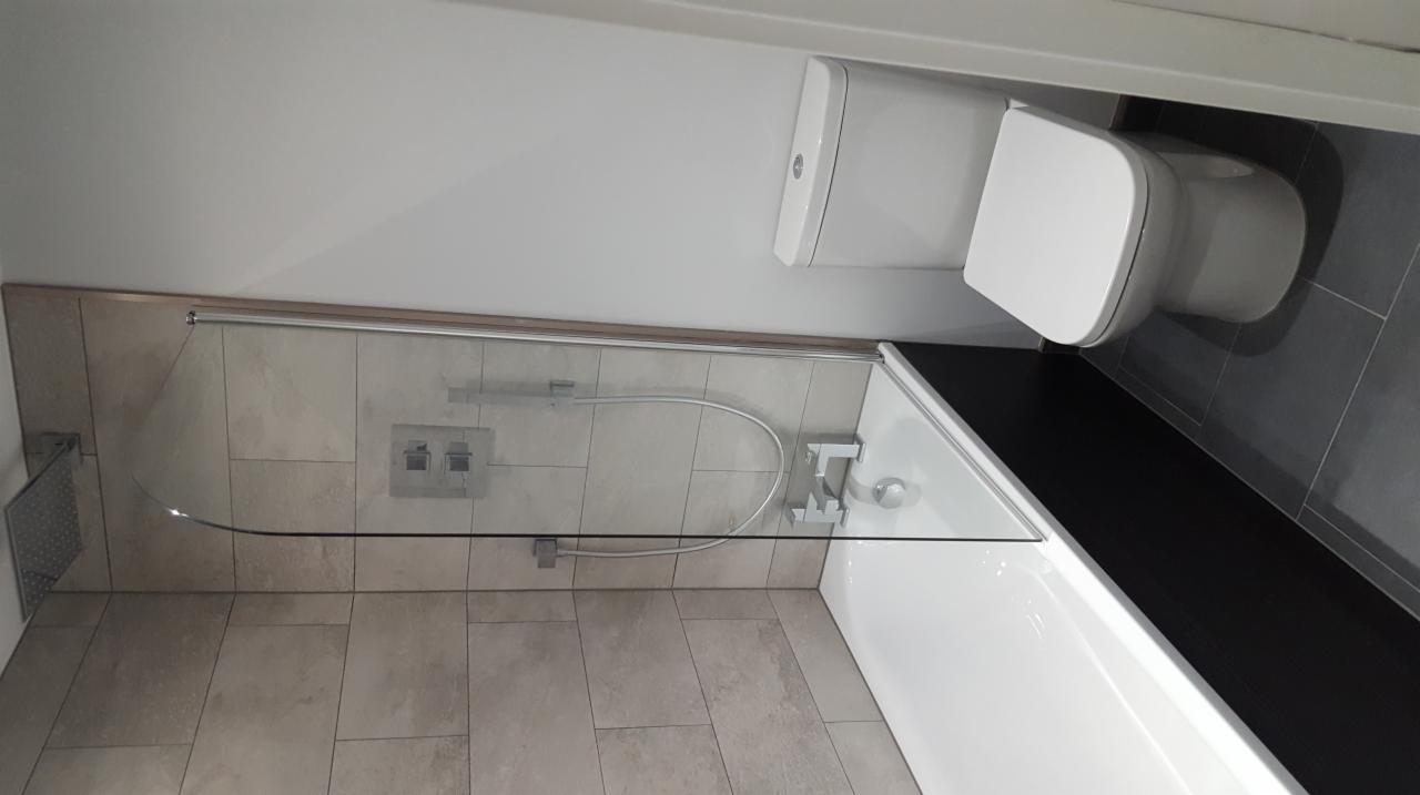 Nowoczesna łazienka po remoncie: beżowe płytki na ścianach, szklany parawan nawannowy z chromowanymi elementami, biała wanna i toaleta, ciemnoszara podłoga z płytek.