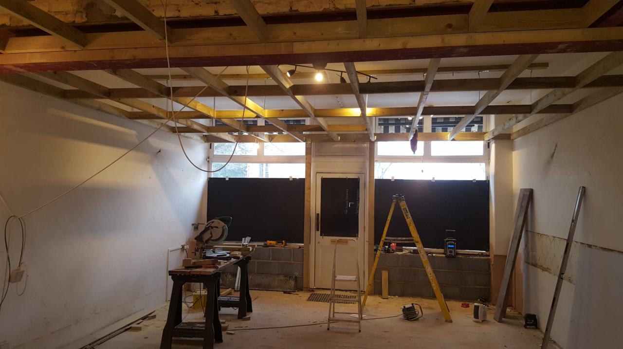 Wnętrze w trakcie remontu: widoczne drewniane elementy konstrukcji sufitu, ściany przygotowane do prac wykończeniowych, narzędzia i drabina w pomieszczeniu.