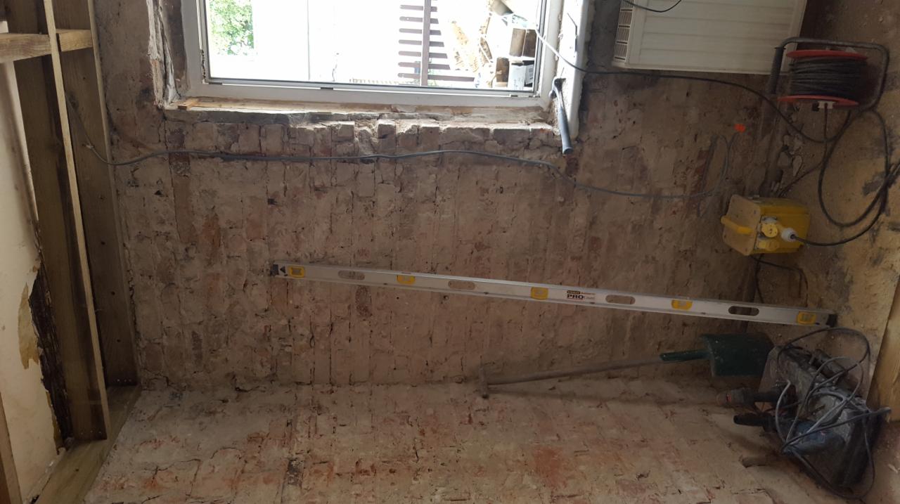 Surowa ściana z czerwonej cegły w trakcie remontu, widoczna poziomica, przewody elektryczne, okno w górnej części kadru i częściowo otynkowana ściana po lewej stronie.