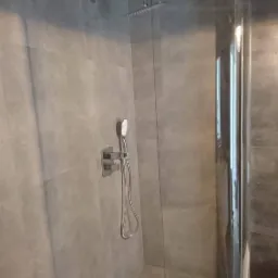 Szara łazienka z kabiną prysznicową typu walk-in, deszczownicą i baterią podtynkową, wykończona płytkami imitującymi beton.