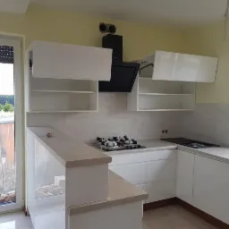 Nowoczesna, biała kuchnia na wymiar z otwartymi szafkami wiszącymi i wyspą, widok na zewnątrz przez okno balkonowe, płytki imitujące cegłę na ścianie.