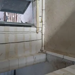 Surowe wnętrze w trakcie prac: kable elektryczne ułożone w bruzdach w ścianie z białych bloczków, widoczne pomarańczowe puszki instalacyjne oraz pionowe prowadzenie kabli przy ścianie.