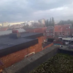 Widok z dachu na zabudowę miejską w pochmurny dzień, z widoczną betonową attyką porośniętą mchem na pierwszym planie.