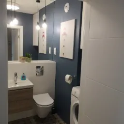 Nowoczesna łazienka z ciemnoniebieską ścianą, wzorzystą podłogą, umywalką z drewnianą szafką, toaletą, pralką Electrolux i obrazami latarni morskich.