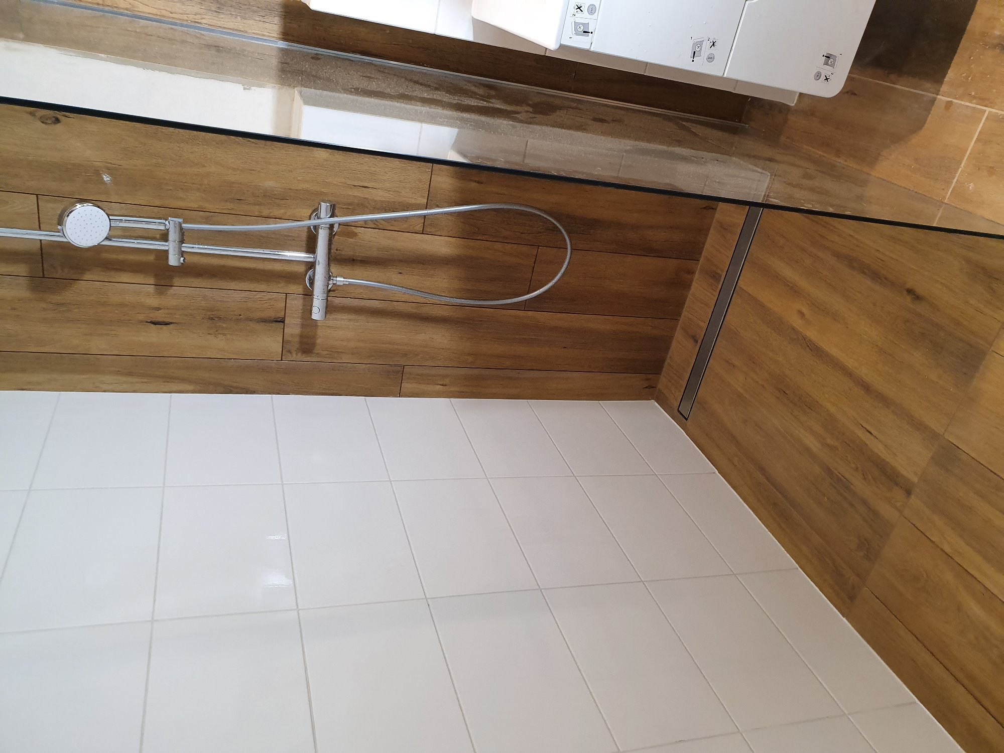 Nowoczesna kabina prysznicowa z drewnopodobnymi płytkami ściennymi i białymi płytkami podłogowymi, widoczny odpływ liniowy i chromowana bateria.