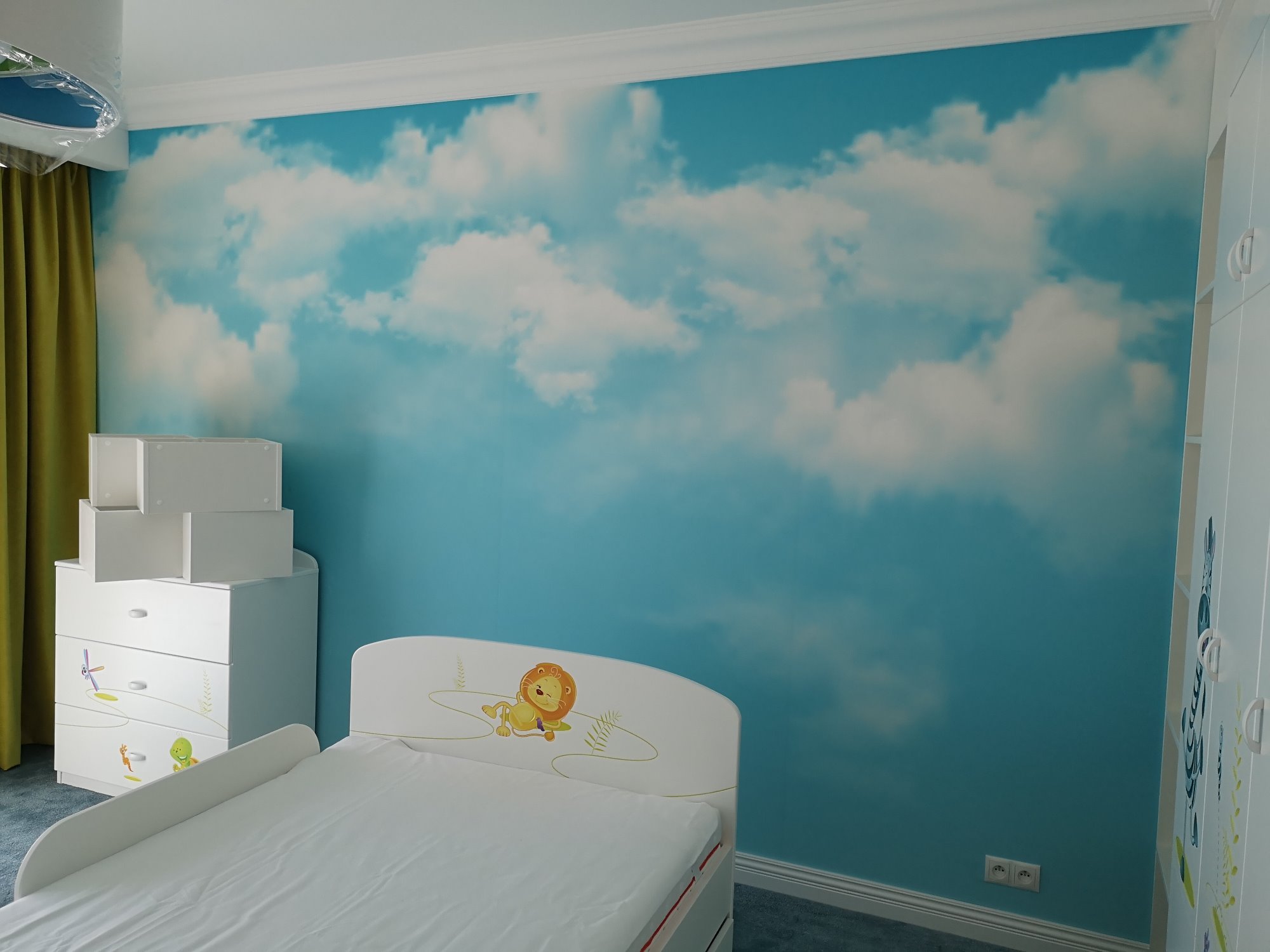 Pokój dziecięcy z fototapetą przedstawiającą niebo z chmurami, białe meble z motywami zwierzęcymi: łóżko z lwem, komoda z wiatrakiem i dinozaurem, oraz szafa z podłużnymi uchwytami.