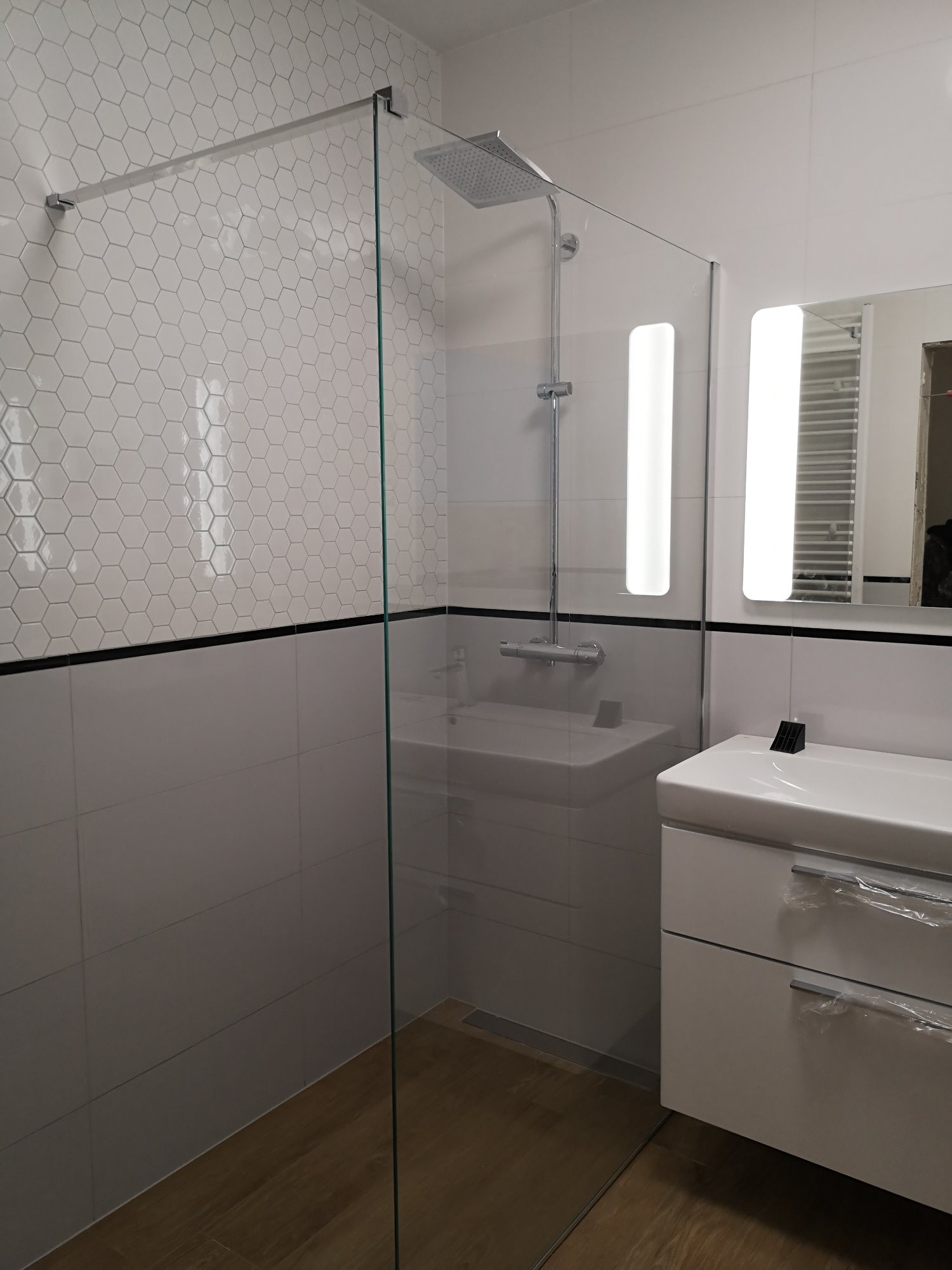 Nowoczesna łazienka z kabiną prysznicową typu walk-in, heksagonalnymi płytkami na ścianie i szafką pod umywalką z folią ochronną na uchwytach szuflad.