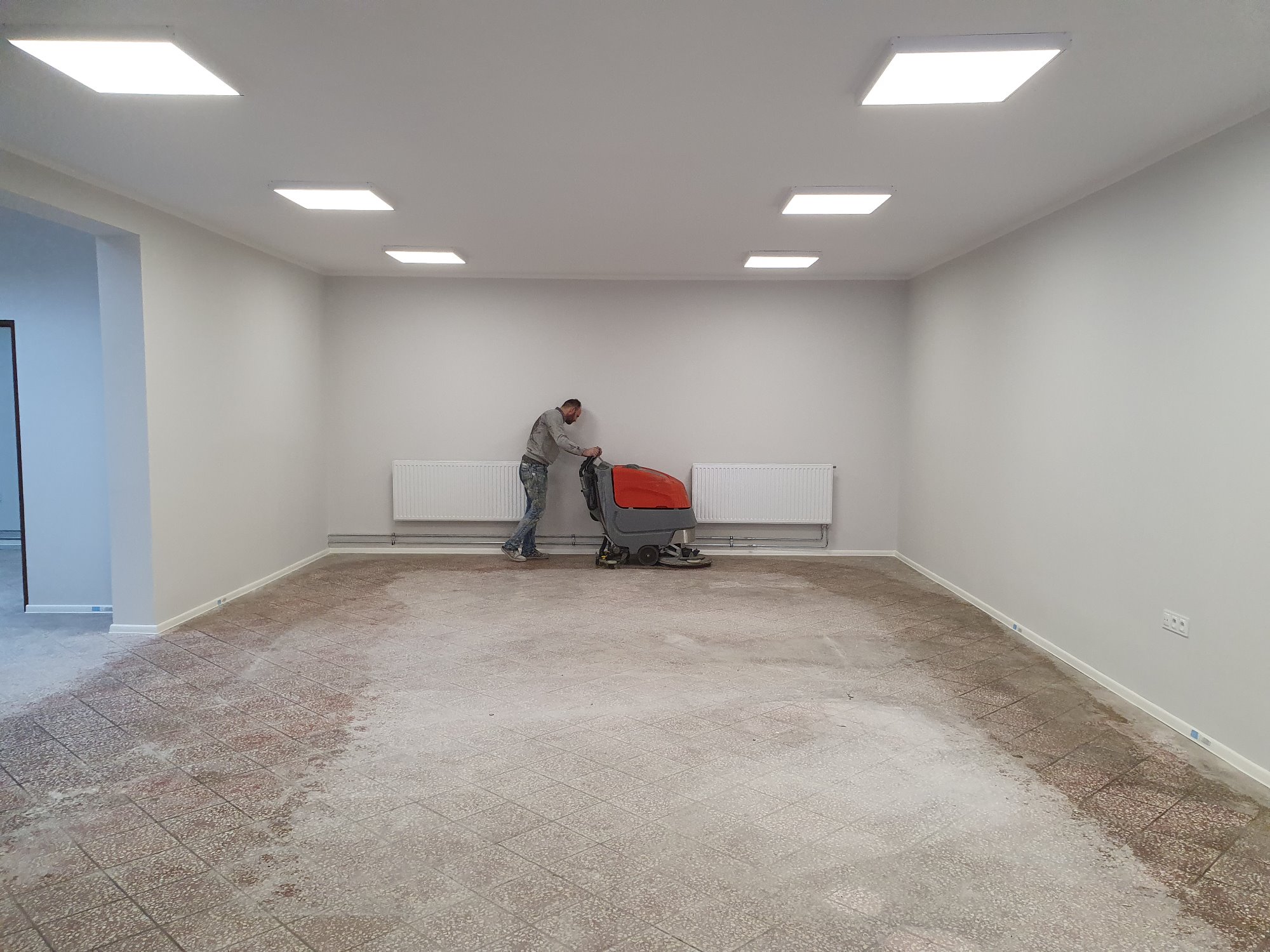 Czyszczenie podłogi w pomieszczeniu za pomocą profesjonalnej maszyny czyszczącej; widoczne ślady po czyszczeniu na kafelkach podłogowych w przestronnym pokoju z grzejnikami.