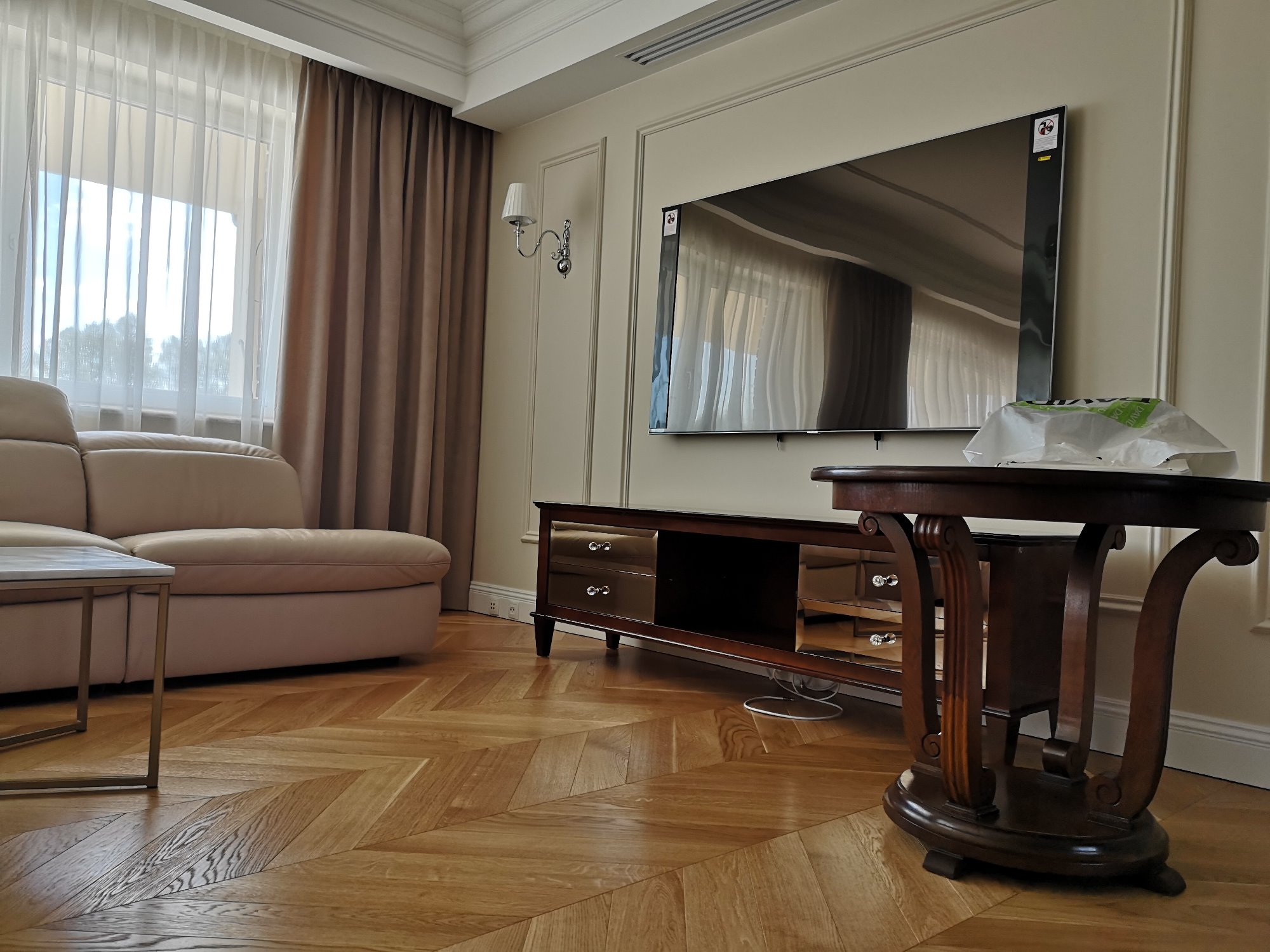 Salon z parkietem w jodełkę, beżową sofą, ciemną szafką RTV z kryształowymi gałkami i okrągłym stolikiem z dekoracyjnymi nogami.