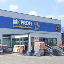 Fasada budynku z szyldem 'psb (PROFI) materiały budowlane', wystawka paneli podłogowych i palety z workami przed wejściem.
