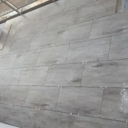 Układanie podłużnych, szarych płytek imitujących drewno na zewnątrz budynku, z użyciem krzyżyków dystansowych, widoczne ślady białego pyłu.