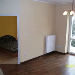 Pusty pokój po remoncie z ciemną podłogą, beżowymi ścianami, białym grzejnikiem i widokiem na zaśnieżony balkon przez okno; w tle widoczny pokój z żółtymi ścianami i wzorzystym łóżkiem.