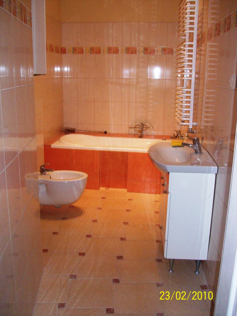 Wykończona łazienka w Bydgoszczy z beżowymi płytkami podłogowymi z brązowymi akcentami, białą wanną w obudowie, bidetem i umywalką z szafką oraz grzejnikiem drabinkowym.