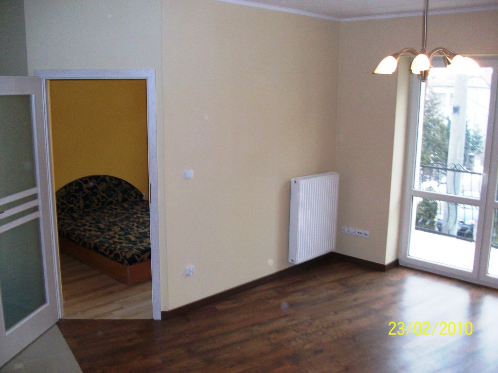 Pusty pokój po remoncie z ciemną podłogą, beżowymi ścianami, białym grzejnikiem i widokiem na zaśnieżony balkon przez okno; w tle widoczny pokój z żółtymi ścianami i wzorzystym łóżkiem.