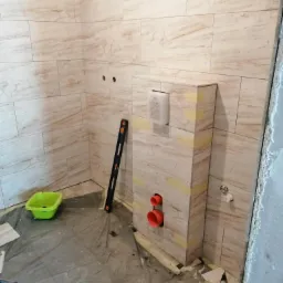 Łazienka w trakcie wykańczania: obudowa stelaża WC z płytkami imitującymi marmur, widoczne pomarańczowe rury, poziomica oparta o ścianę, zabezpieczona podłoga i fragmenty płytek.