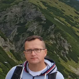 Mężczyzna w okularach i bluzie z kapturem stoi na tle górskiego krajobrazu z widocznymi skałami i zieloną roślinnością na zboczach.