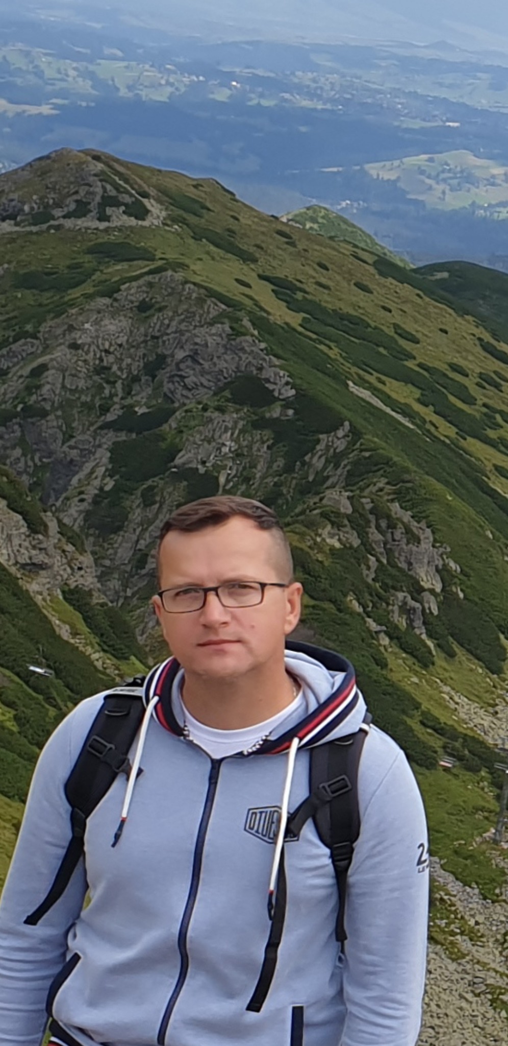 Mężczyzna w okularach i bluzie z kapturem stoi na tle górskiego krajobrazu z widocznymi skałami i zieloną roślinnością na zboczach.