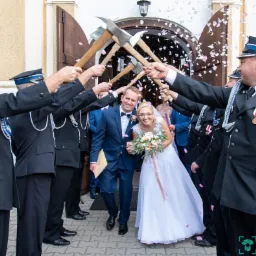 Para młoda wychodzi z kościoła pod szpalerem strażaków z toporami, konfetti w powietrzu, uśmiechnięci.