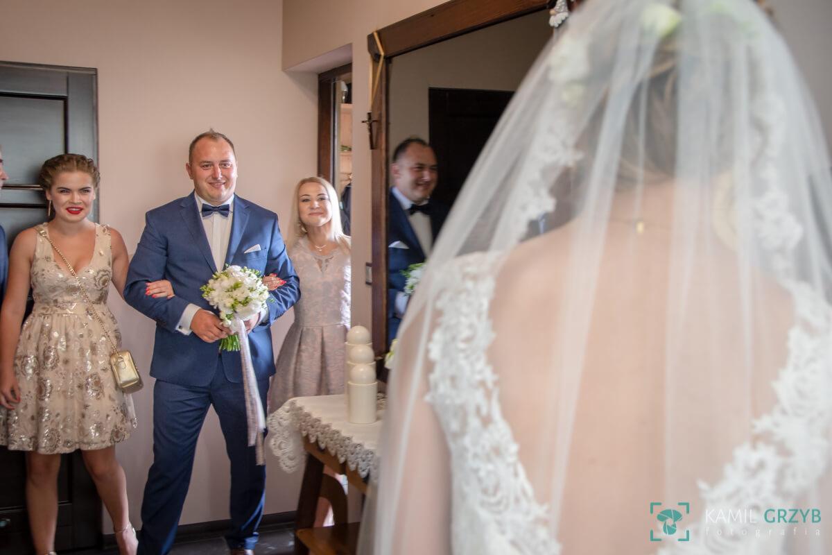 Panna młoda w welonie, widziana od tyłu, oczekująca na wejście, z widocznym panem młodym prowadzonym przez dwie kobiety, w tle lustrzane odbicie pana młodego.