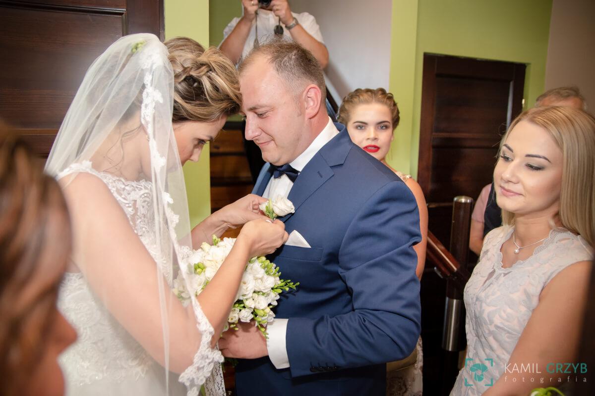 Panna młoda przypina butonierkę z białych kwiatów do klapy granatowego garnituru pana młodego, w tle goście weselni i fotograf.