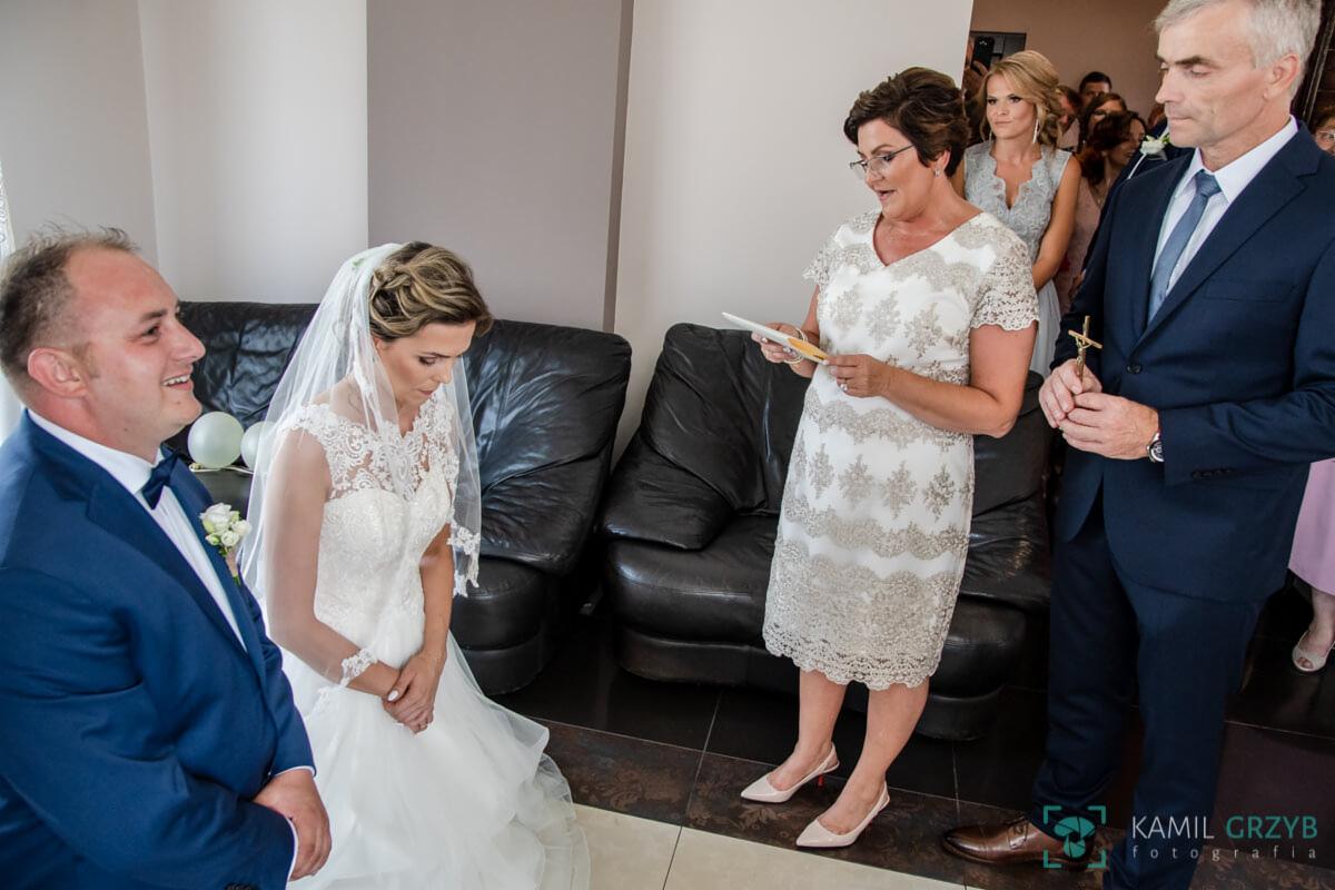 Panna młoda w koronkowej sukni klęczy podczas ceremonii, matka czyta tekst, a goście obserwują z tyłu.