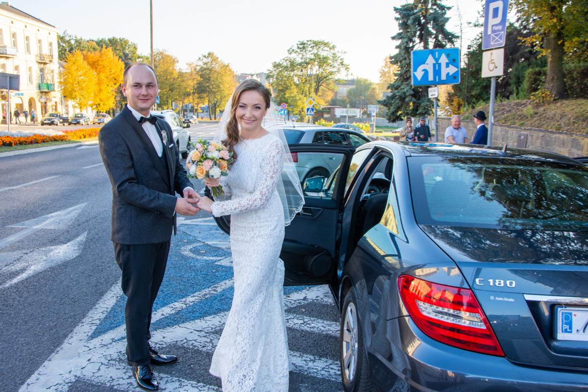 Para młoda trzymająca się za ręce obok otwartych drzwi samochodu Mercedes C180 na tle miejskiego krajobrazu z drzewami o jesiennych barwach i przechodniami.
