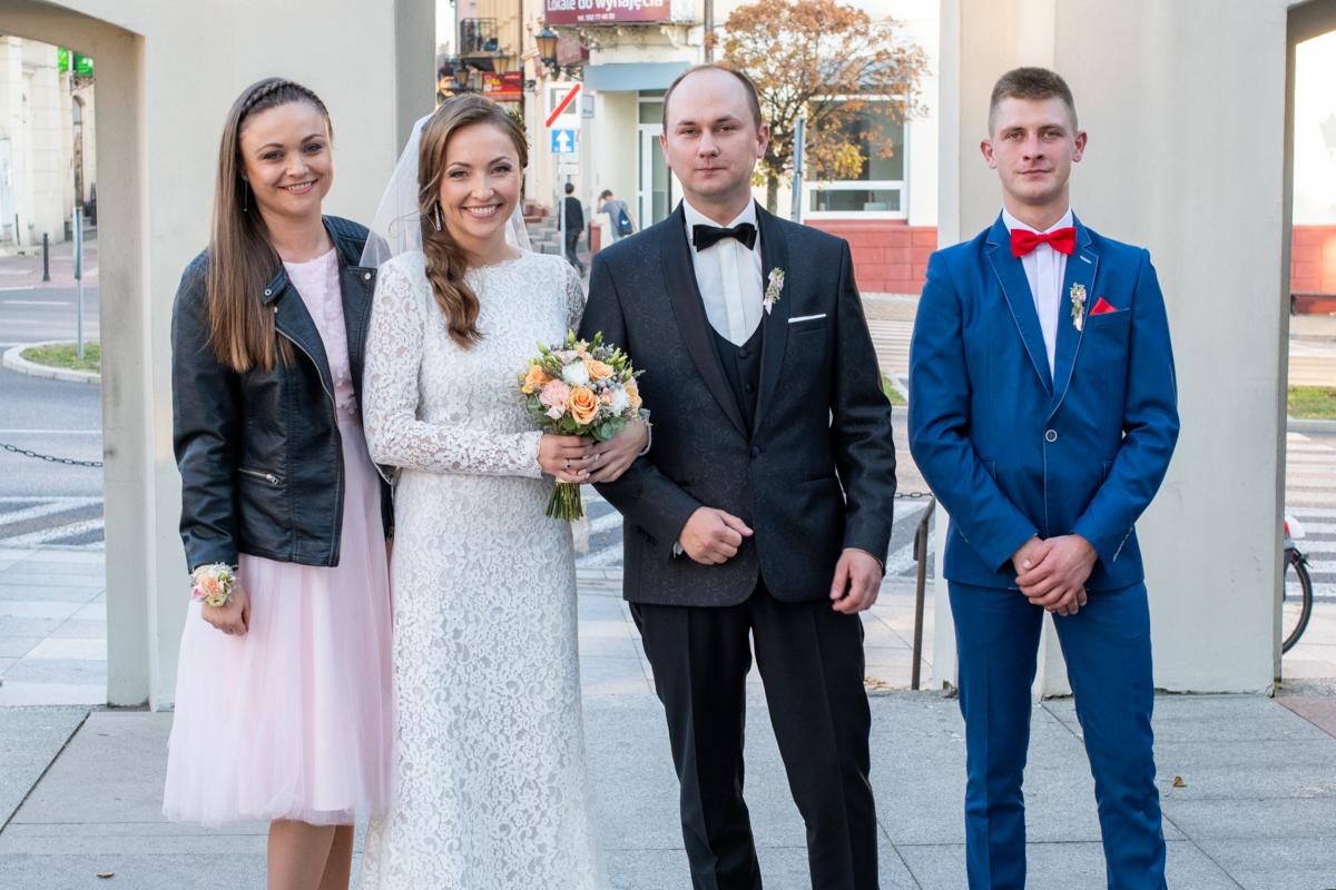 Uśmiechnięta para młoda z bukietem i świadkami pozuje do zdjęcia na tle miejskiego placu z budynkami i drzewami.